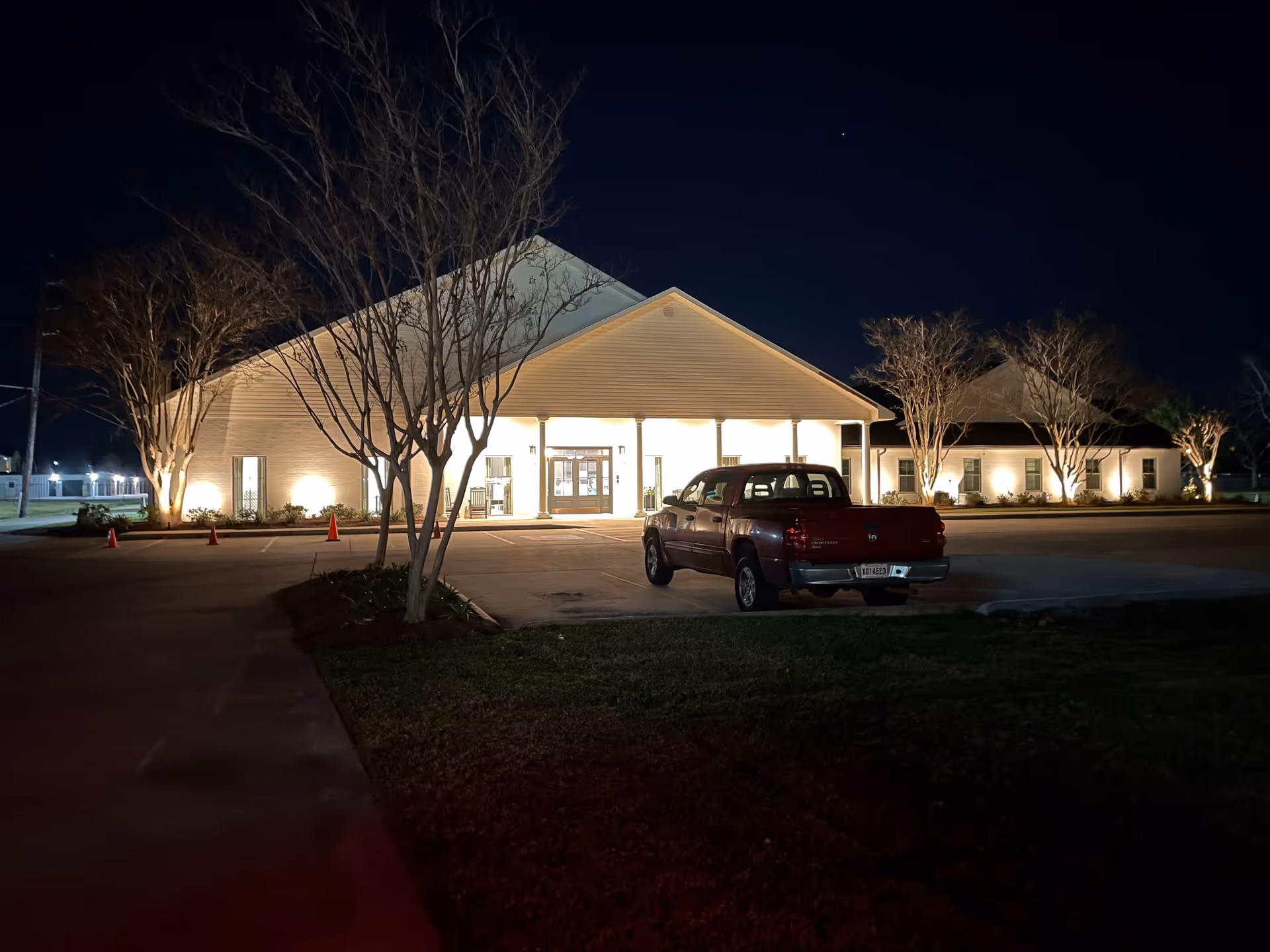 Nighttime view of a well-lit single-story building with a peaked roof, surrounded by leafless trees and a parking lot with a red pickup truck parked near the entrance.