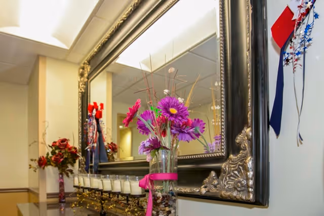 A decorative interior scene featuring a large ornate mirror mounted on a wall above a surface with a vase of purple flowers and a row of small candles. There are additional floral decorations and patriotic ribbons on the wall nearby.