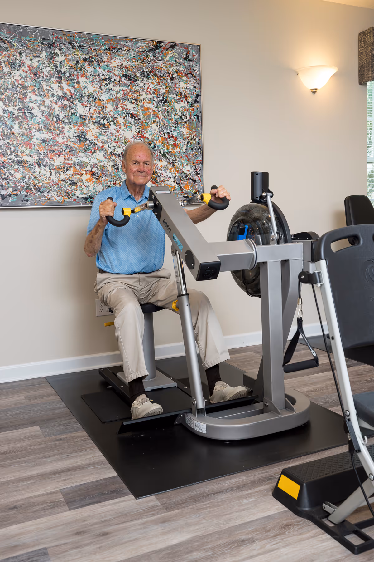 An elderly man using seated exercise equipment in a fitness room with abstract wall art on the wall.