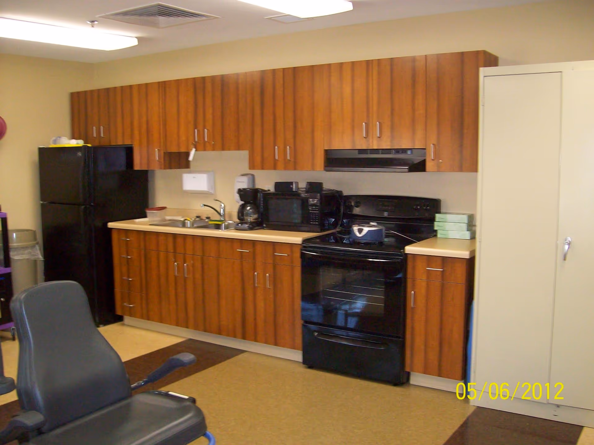 Interior view of a kitchen area with wooden cabinets, a black refrigerator, a black stove with oven, a microwave, a coffee maker, a sink, and a beige countertop. There is a gray chair in the foreground and a white storage cabinet on the right side.
