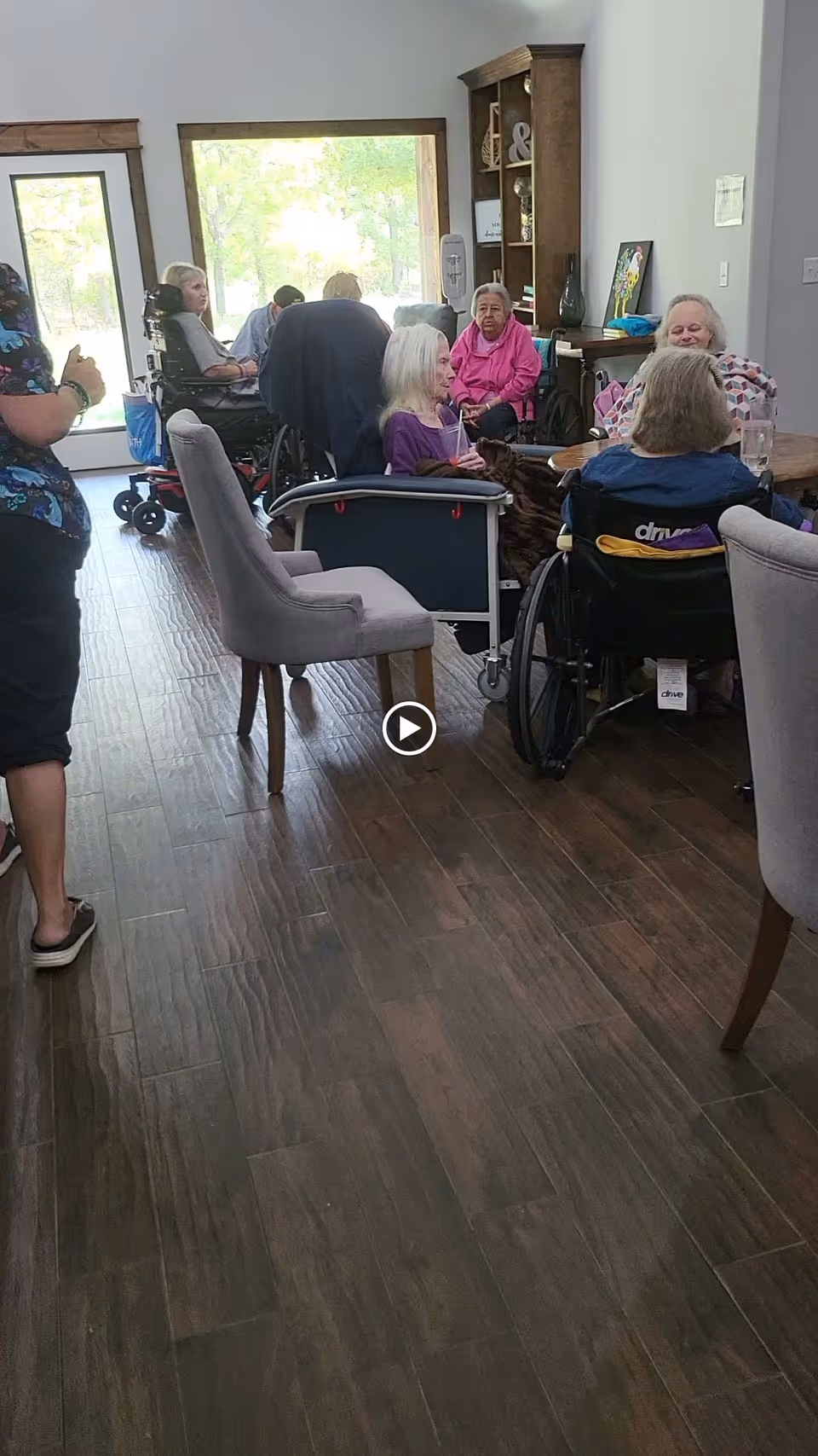 Several elderly residents in wheelchairs and staff gathered in a bright common area with wood flooring and tables.