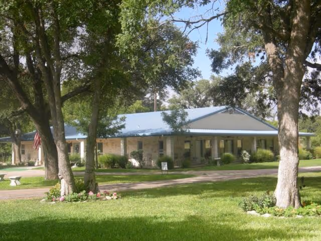 Single-story building with a blue roof surrounded by large trees and green grass under a clear sky, with a pathway leading to the entrance.