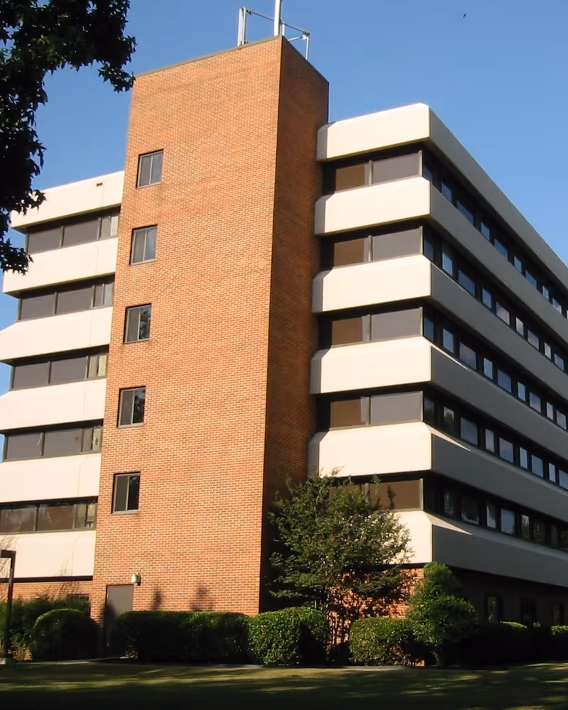 Exterior view of a multi-story building with a brick central section and white horizontal bands with windows on the sides, surrounded by green bushes and trees under a clear blue sky.