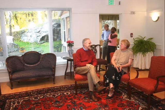 A cozy living room area in a senior living facility with two elderly people sitting on red chairs having a conversation. In the background, a man and a woman are standing near a doorway. The room has a large window showing a green outdoor area, a dark wooden loveseat, a side table with a vase of flowers, a large red patterned rug, and a potted fern on a table covered with a white cloth.