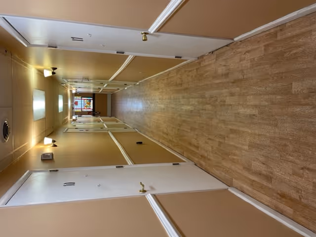 A long indoor hallway with light brown walls and wood-patterned flooring. White doors with numbers 10 and 11 are visible on the left and right sides respectively. The hallway is lit by ceiling lights and wall sconces, and there is a stained glass window at the far end.