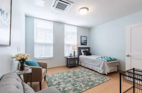 A bright and cozy bedroom with light blue walls, a single bed with gray bedding and a teal throw blanket, a nightstand with a lamp and picture frame, two large windows with white blinds, a patterned area rug, a beige armchair with a teal pillow, a gray sofa, a small side table with a vase of white flowers, and a closed white door.
