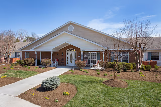 Front entrance of a single-story brick and siding senior living building with a covered entry, walkway, rocking chairs, and landscaped lawn.