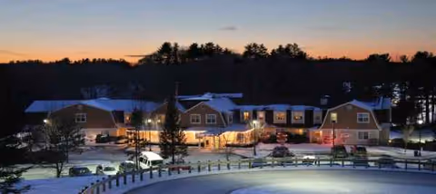Snow-covered senior living campus at dusk with warmly lit entrance and parked cars.