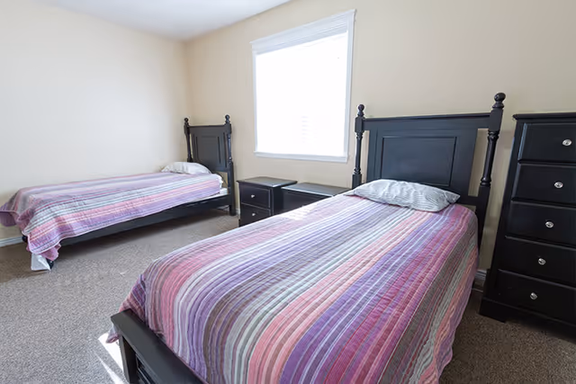 Two twin beds with striped purple bedspreads in a bright bedroom with black wooden headboards, nightstands, and a window.