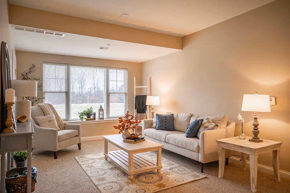 A cozy, well-lit living room with a sofa, armchair, coffee table, and a large window overlooking a lawn.