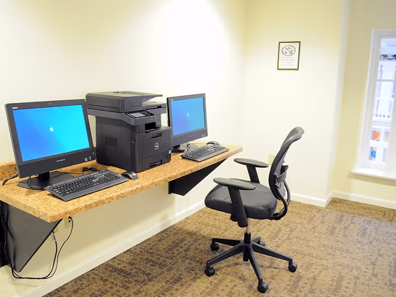 A small computer workstation area with two desktop computers, a printer placed between them on a wall-mounted wooden desk. There is a black office chair on wheels in front of the desk. The room has beige walls, a carpeted floor, and a window on the right side letting in natural light.