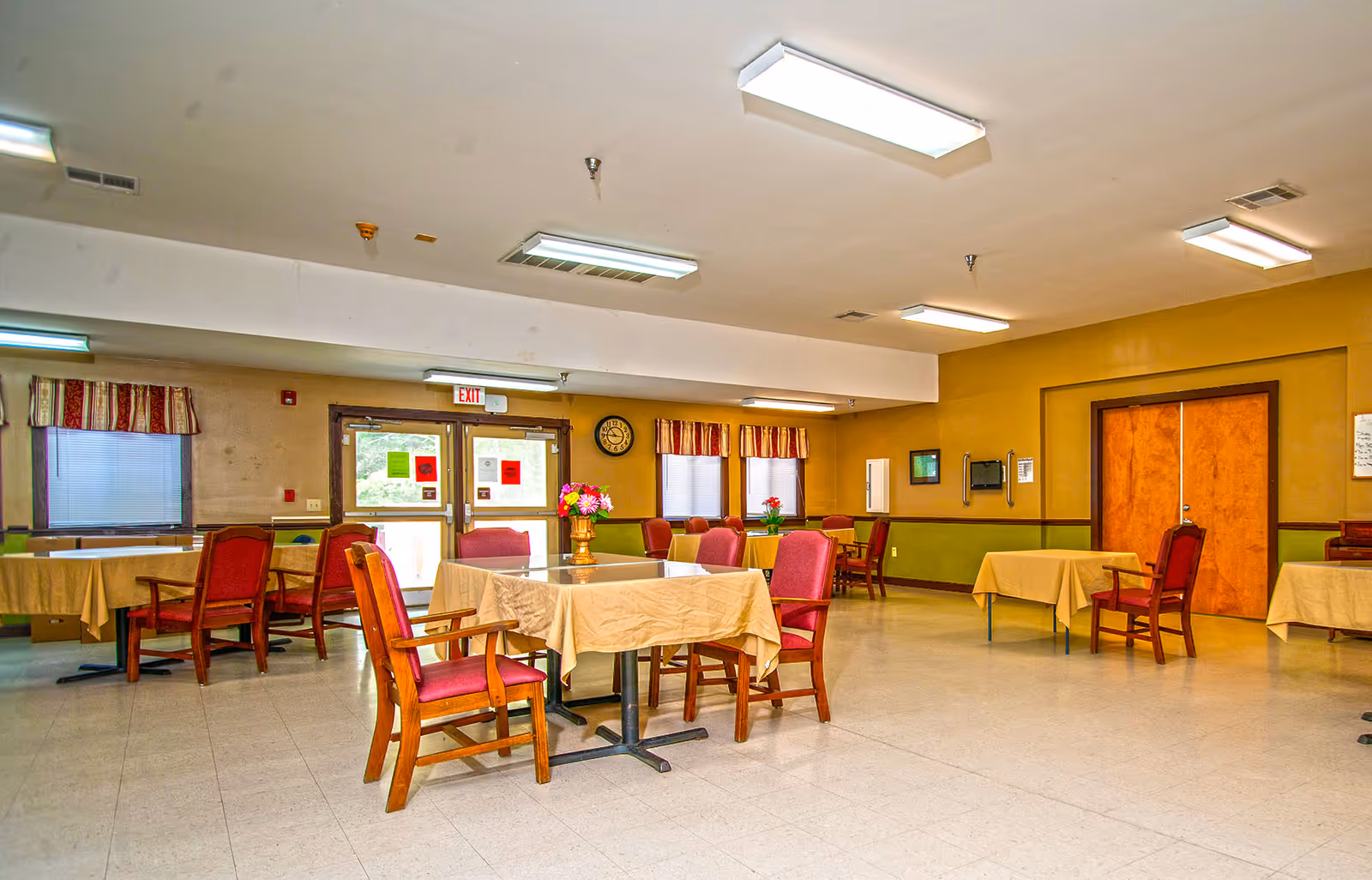 A spacious dining room with several tables covered in beige tablecloths and surrounded by wooden chairs with red cushions. The room has yellow and green walls, fluorescent ceiling lights, windows with striped valances, and a clock on the wall. There are double doors with an exit sign above them and flower arrangements on some tables.