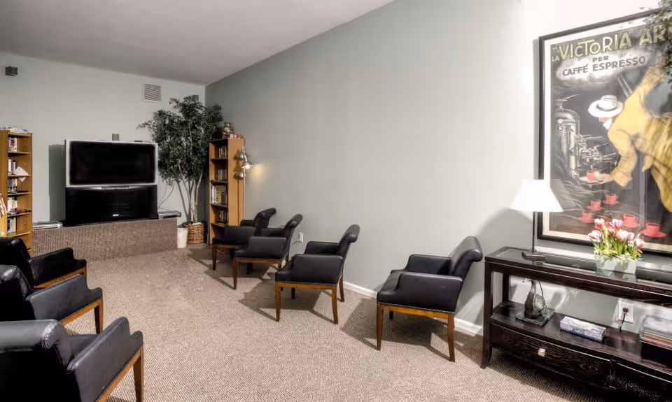 A cozy living room with six black armchairs arranged facing a television on a stand. There are two wooden bookshelves on either side of the TV, a tall potted plant in the corner, and a framed vintage coffee poster on the wall above a dark wooden console table with a lamp and a vase of flowers.