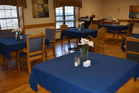 Dining room with blue-clothed tables and wooden chairs arranged around a serving counter, each table decorated with a small vase of flowers.