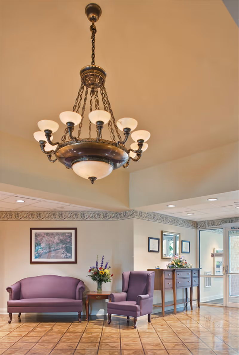 A well-lit seating area in a senior living facility featuring a purple loveseat and matching armchair, a wooden side table with a vase of flowers, a wooden console table with floral arrangements, framed artwork and certificates on the walls, a large ornate chandelier hanging from the ceiling, and a glass door entrance in the background.