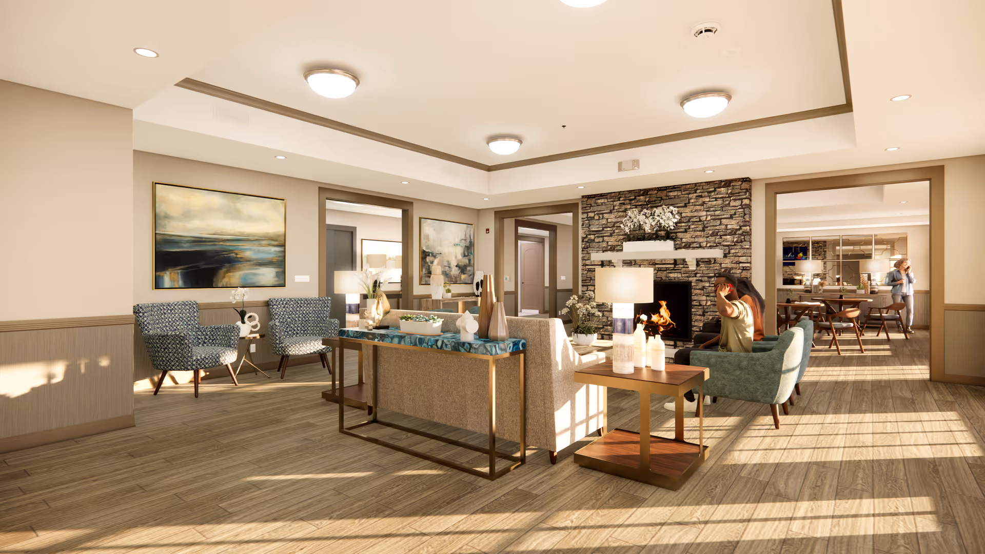 A bright and spacious senior living common area with wooden flooring, modern furniture including armchairs and sofas arranged around a stone fireplace with a fire burning. The room features large paintings on the walls, decorative lamps, and a table with vases and plants. In the background, there is a dining area with a person standing near the table.
