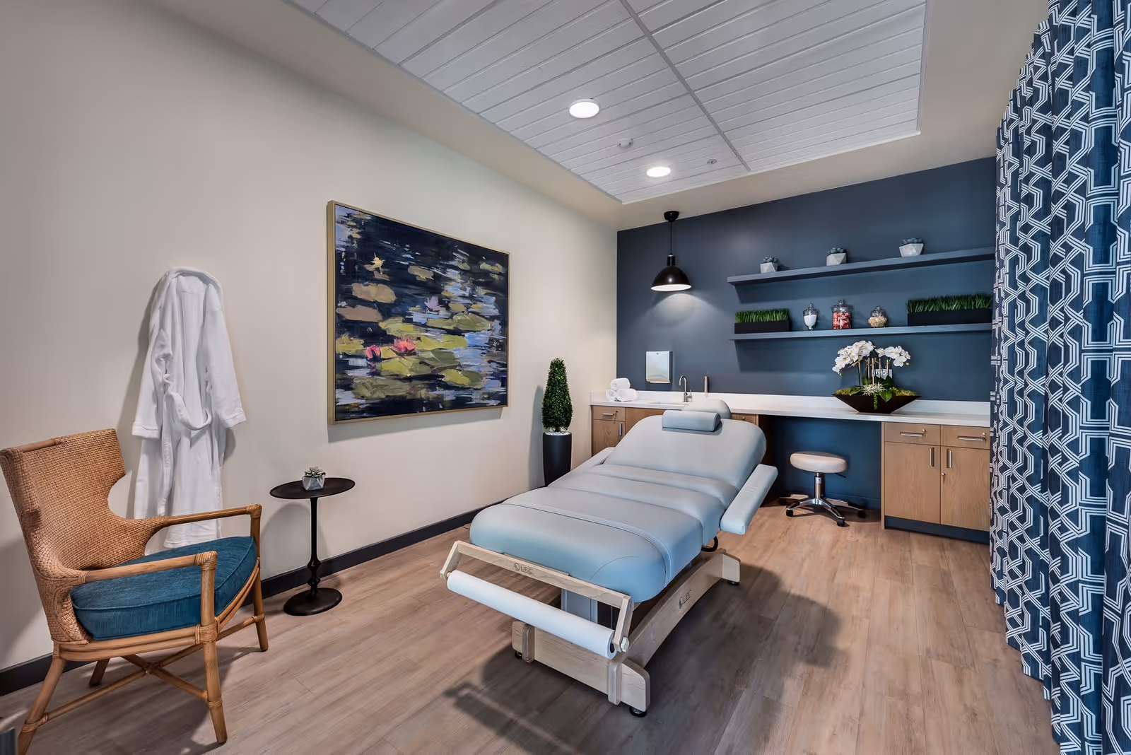 A modern treatment room with a padded reclining medical or massage table in the center. The room features a wicker chair with a blue cushion, a small round side table, a white robe hanging on the wall, and a large abstract painting. The back wall has wooden cabinets, open shelves with decorative plants and jars, a sink, and a black pendant light. A blue patterned curtain is partially visible on the right side.