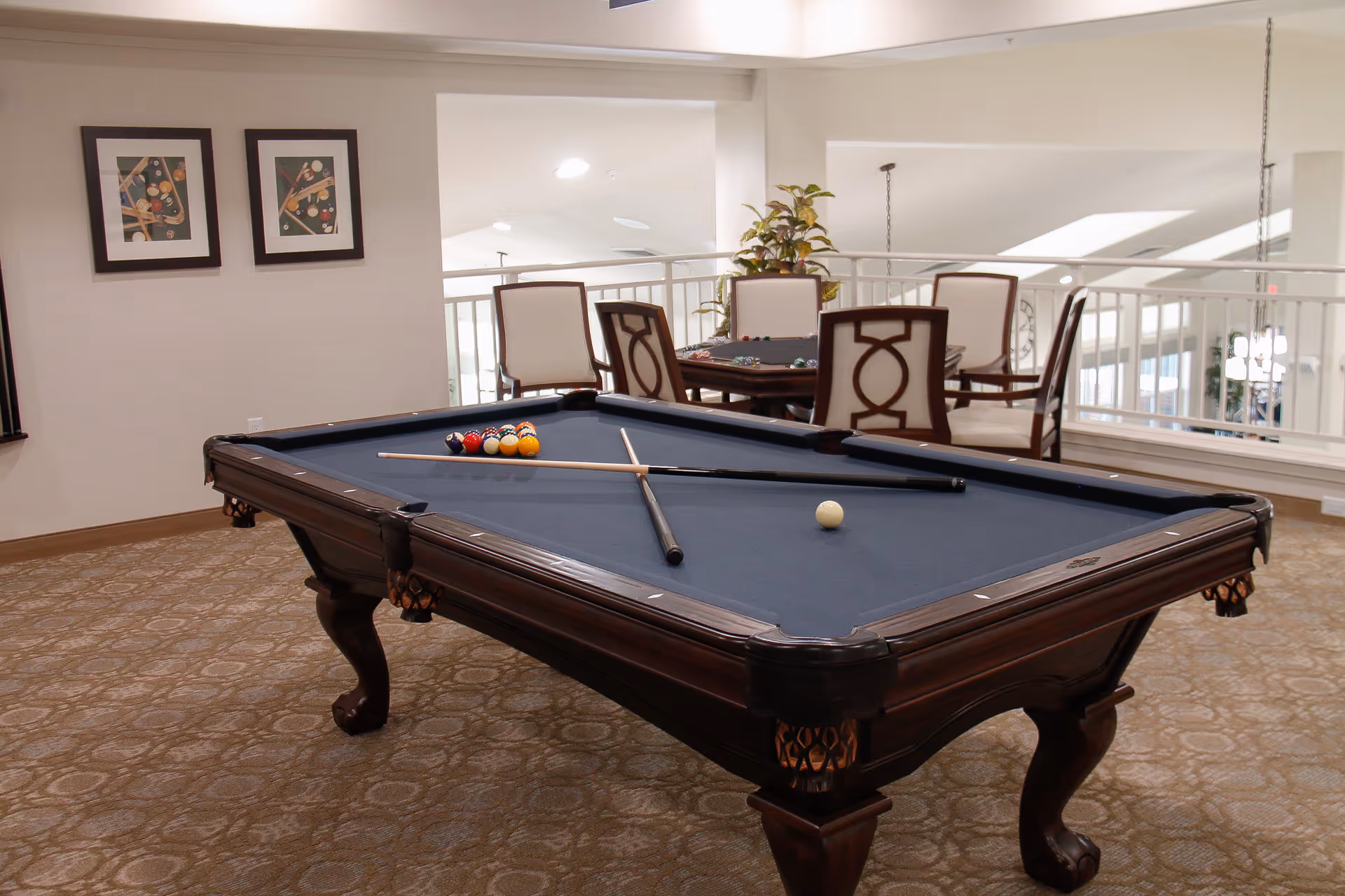 A blue-felt pool table with cues and racked balls in a well-lit lounge area with chairs and framed artwork on the wall.