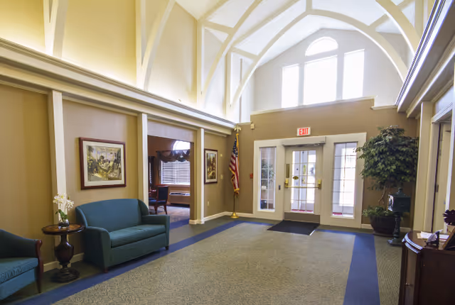 A bright assisted-living lobby with a vaulted ceiling, seating, potted plants, and double glass entrance doors with an EXIT sign.
