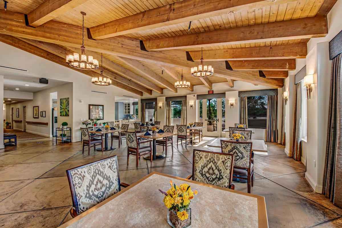 A spacious dining room with wooden beams on the ceiling and multiple chandeliers. Several tables with patterned chairs are arranged throughout the room, each table decorated with a small flower arrangement. Large windows and glass doors allow natural light to fill the space, and curtains frame the windows. The floor has a polished concrete finish with a geometric pattern.