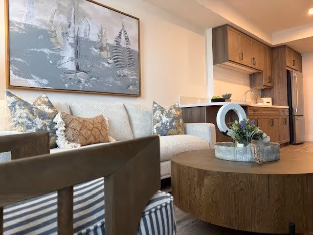 A modern living room with a beige sofa adorned with decorative pillows, a round wooden coffee table with a floral arrangement, and a wooden chair with striped upholstery in the foreground. In the background, there is a kitchen area with wooden cabinets, a refrigerator, and a countertop. A large framed artwork depicting sailboats hangs on the wall above the sofa.