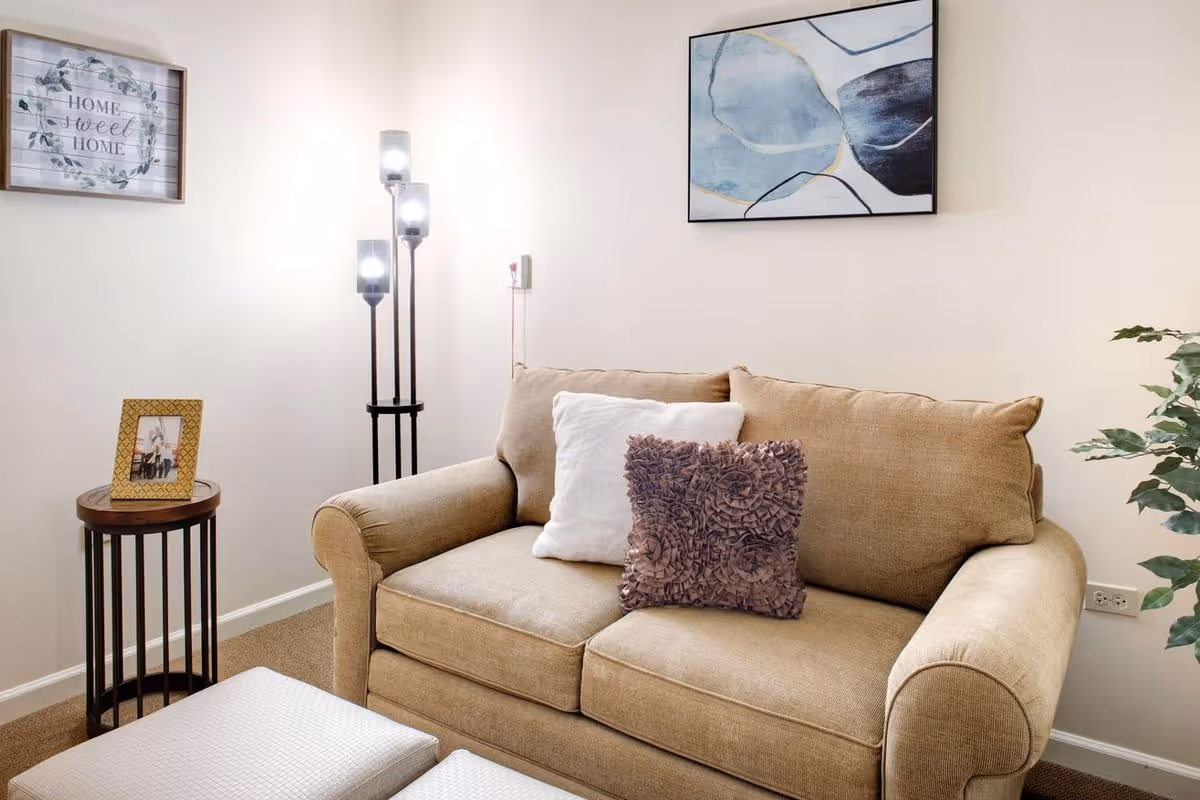 Cozy living room with a beige loveseat, decorative pillows, a floor lamp, side table with a framed photo, wall art, and a potted plant.
