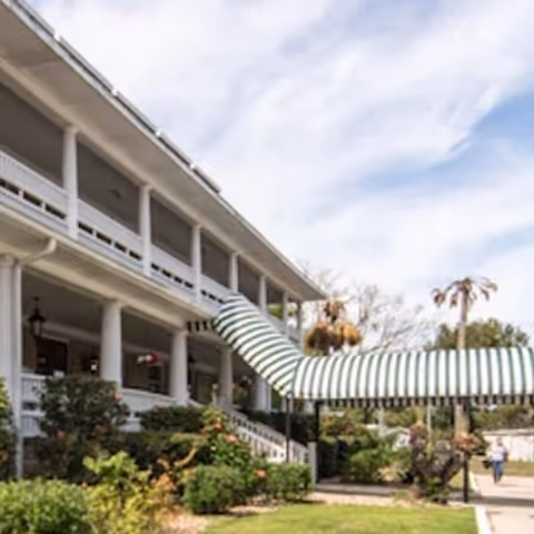 Exterior view of a two-story building with white columns and a striped green and white awning extending from the upper balcony to the ground level. There are bushes and plants in the landscaped area in front of the building, and a person walking on the pathway nearby under a partly cloudy sky.