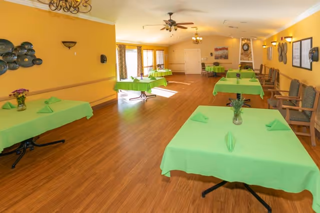 A spacious dining room with several tables covered in bright green tablecloths, each set with folded green napkins and small flower vases. The room has wooden flooring, yellow walls, and multiple chairs lined up against the wall. Large windows on one side allow natural light to fill the room, and ceiling fans and chandeliers provide lighting.