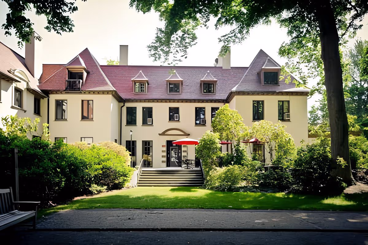 Front exterior of a large cream-colored mansion with a red roof, steps leading to a patio with red umbrellas, and surrounding trees and landscaped lawn.