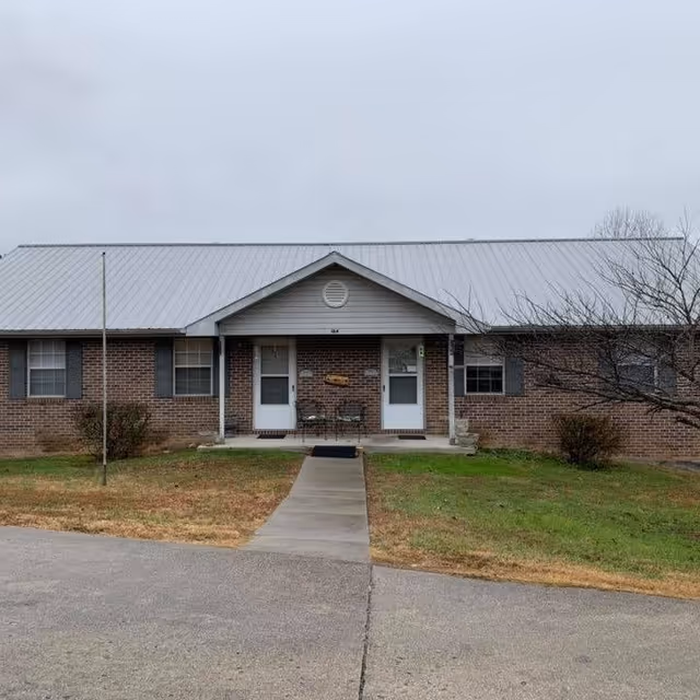 Front exterior view of a single-story brick building with a metal roof, two white doors under a small covered porch, a concrete walkway leading to the entrance, and some grass and bushes on either side.