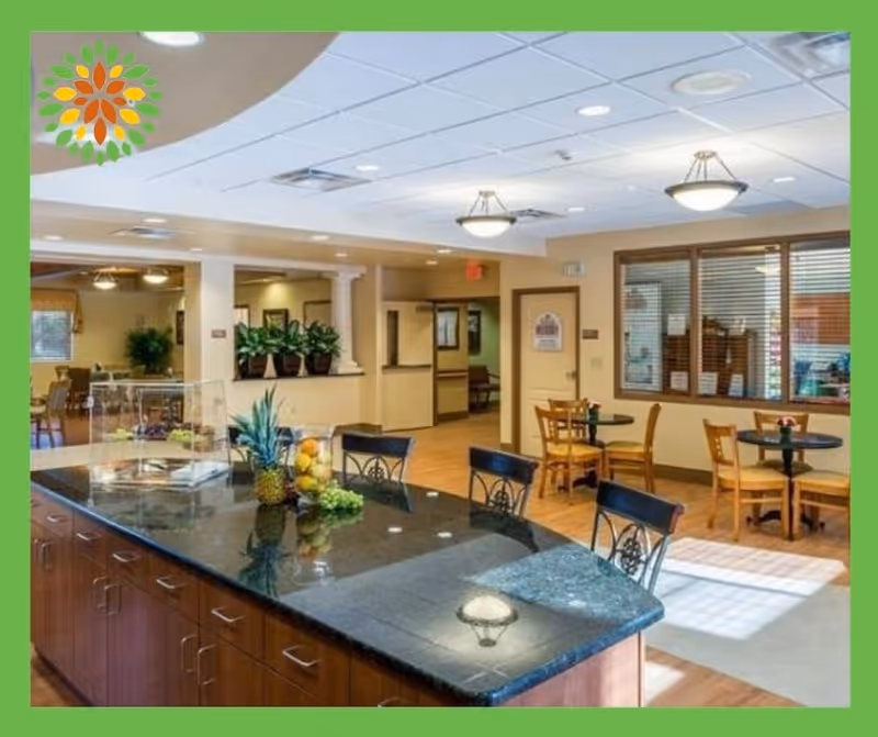 Interior view of a senior living facility dining area with a large black granite countertop island in the foreground decorated with a pineapple, grapes, and other fruits. The background shows multiple small round tables with wooden chairs, potted plants, and a window with blinds. The ceiling has recessed lighting and hanging light fixtures.