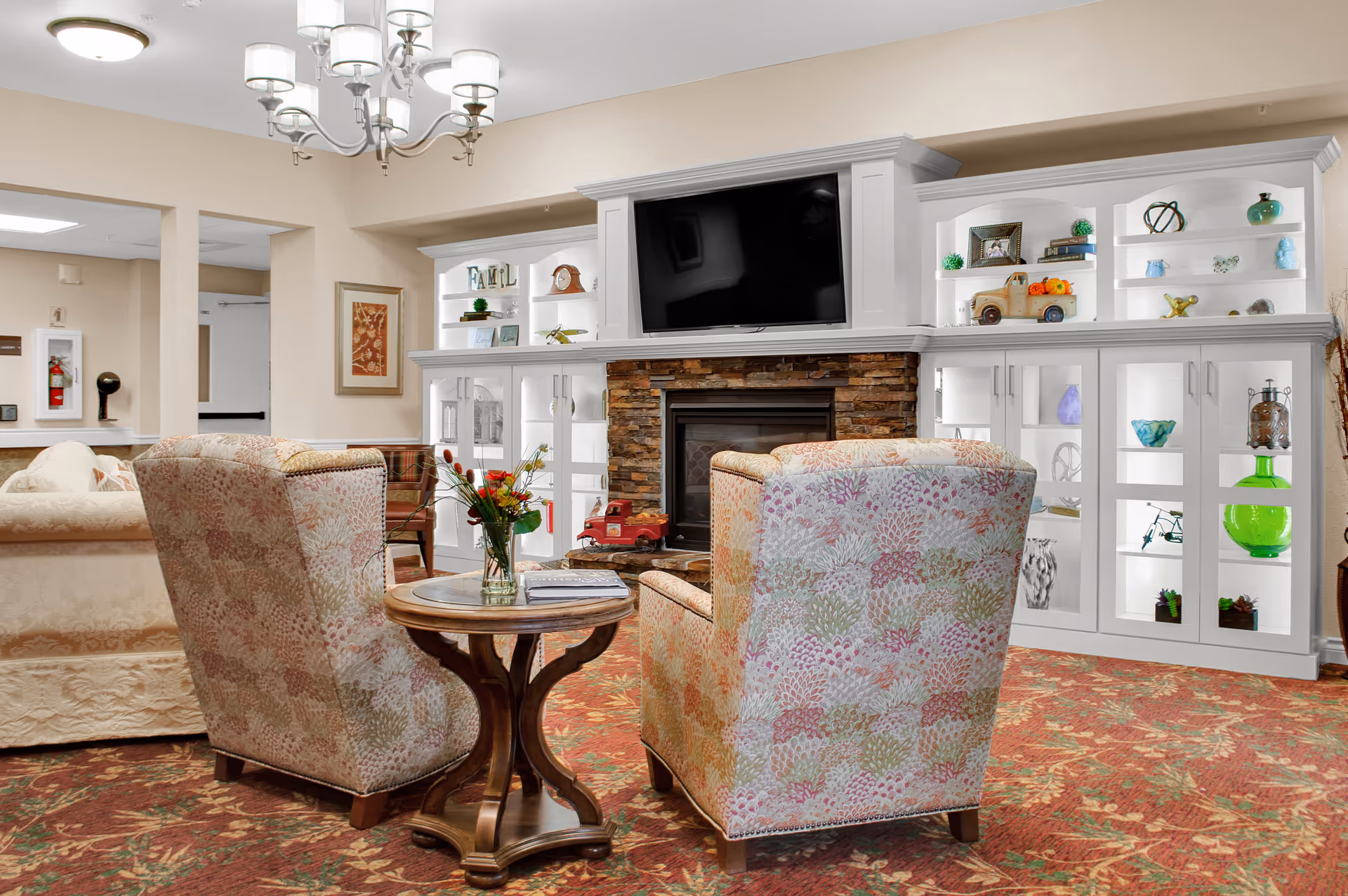 Cozy senior living lounge with patterned armchairs facing a fireplace and wall-mounted TV surrounded by illuminated display cabinets.