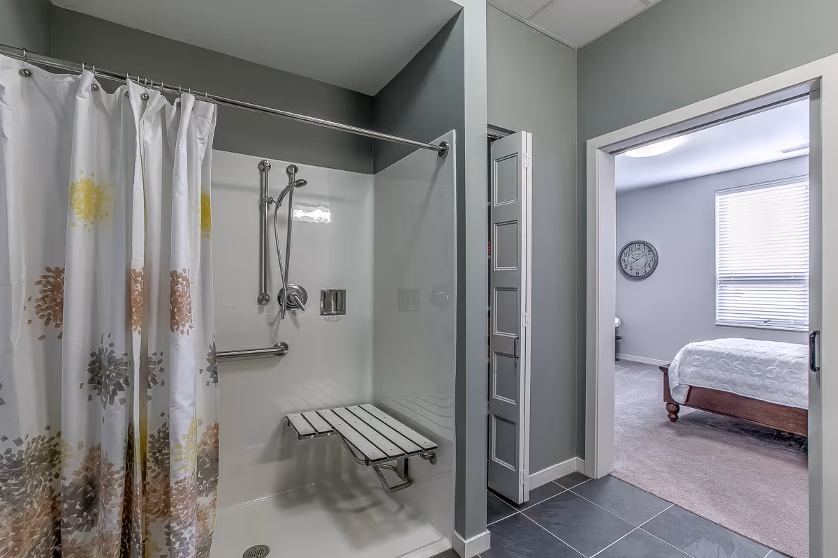 A bathroom with a walk-in shower featuring a white fold-down bench and a shower curtain with a floral pattern in yellow, brown, and gray tones. The shower has a handheld showerhead and grab bars. Adjacent to the bathroom is a bedroom with a bed, a wall clock, and a window with blinds.