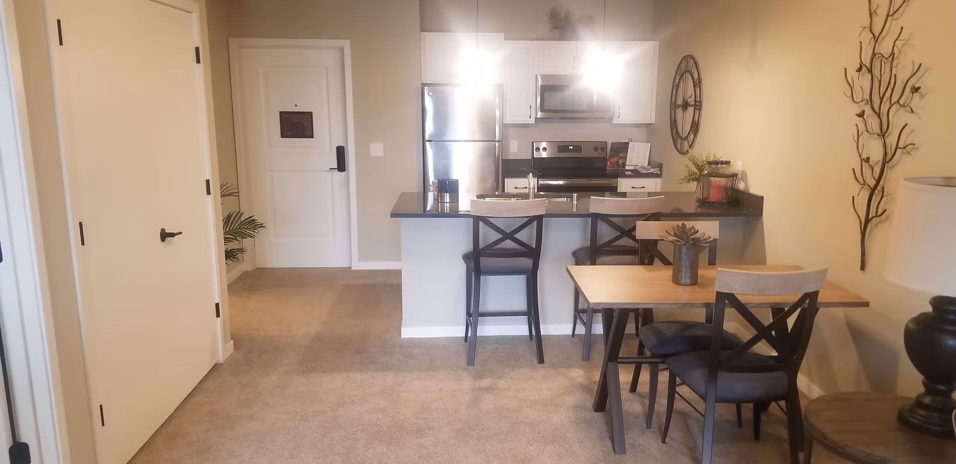Interior view of a senior living facility apartment showing a small kitchen with stainless steel appliances, a breakfast bar with two chairs, and a dining table with three chairs. The room has beige walls, carpeted floor, and decorative elements including a wall clock and a metal wall sculpture.