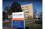 Exterior view of a multi-story building with a large sign in the foreground displaying directions and the name Providence Mount St. Vincent. The sign includes directions to the Main Entrance, West Entrance, Visitor Parking, and St. Joseph Residence. Trees and a clear blue sky are visible in the background.