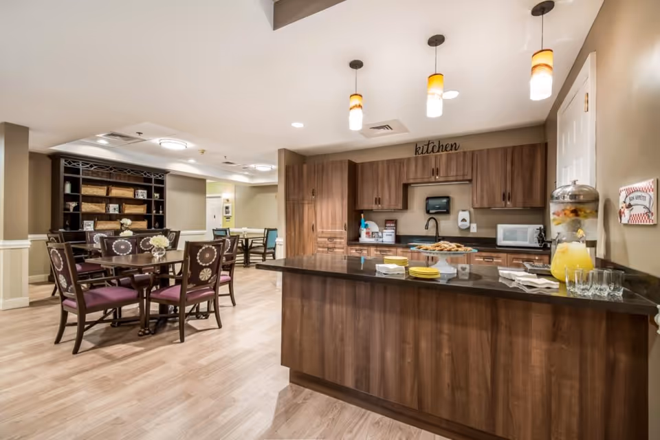 A well-lit communal kitchen and dining area in a senior living facility. The kitchen features wooden cabinets, a black countertop island with plates and a glass dispenser filled with lemonade. There are three pendant lights hanging above the island. In the background, there are dining tables with chairs and a large dark wooden shelving unit with baskets and decorative items.