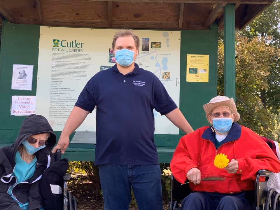 Three people wearing face masks are outdoors in front of a Cutler Botanic Garden information board. Two elderly individuals are seated in wheelchairs on either side of a standing man wearing a navy blue polo shirt with The Homesteads logo. The elderly man on the right is holding a yellow flower and wearing a red jacket and a hat. The elderly woman on the left is wearing a black hooded jacket and glasses.