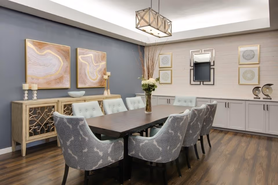 A modern dining room with a long dark wooden table surrounded by eight upholstered chairs with patterned fabric. The room features wooden flooring, a sideboard with decorative items including candles and a bowl, and framed abstract artwork on the walls. A rectangular chandelier hangs above the table, and there is a vase with flowers and tall branches as a centerpiece.