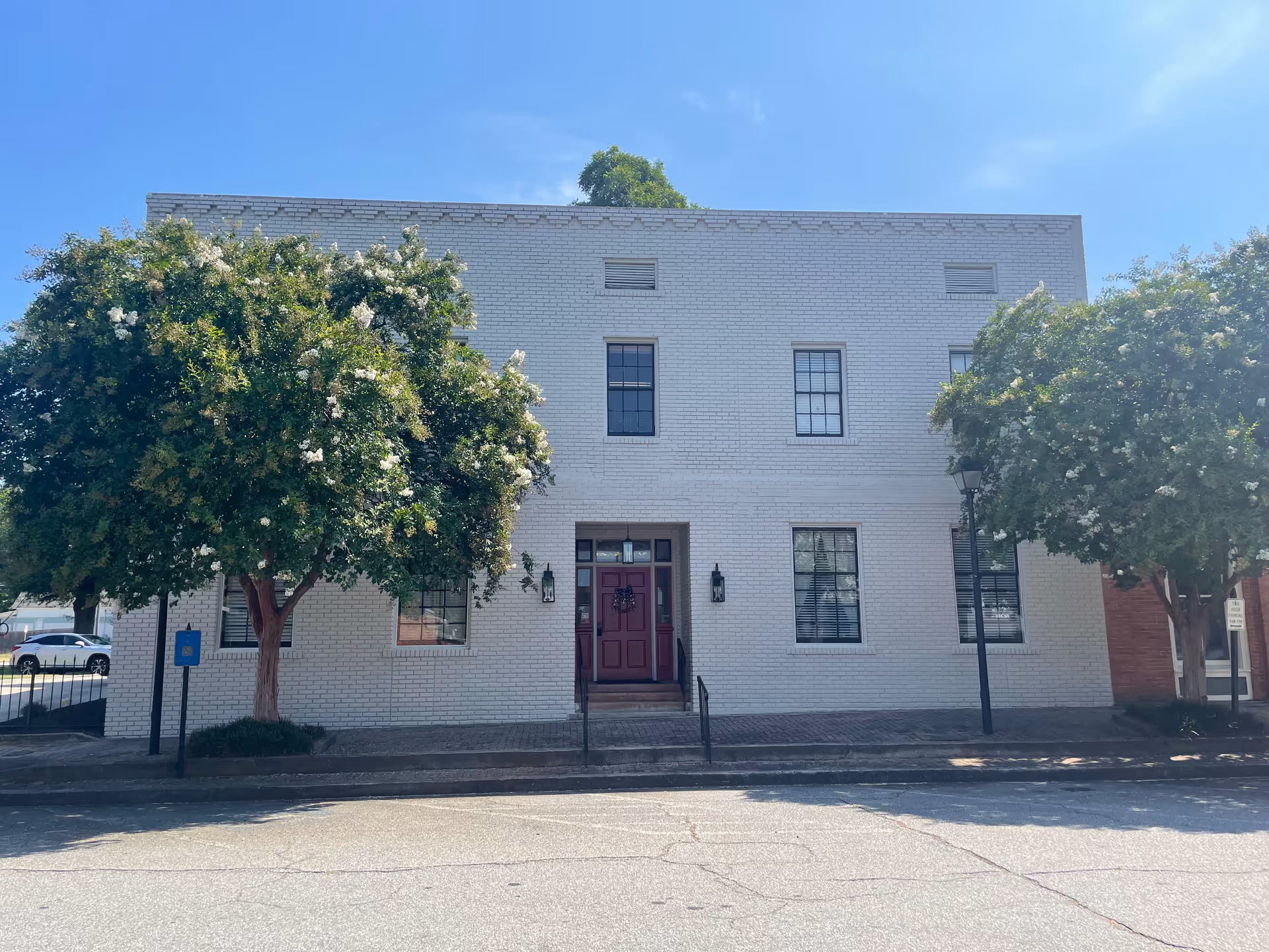 Two-story white brick building with a red entrance door flanked by trees along a street.