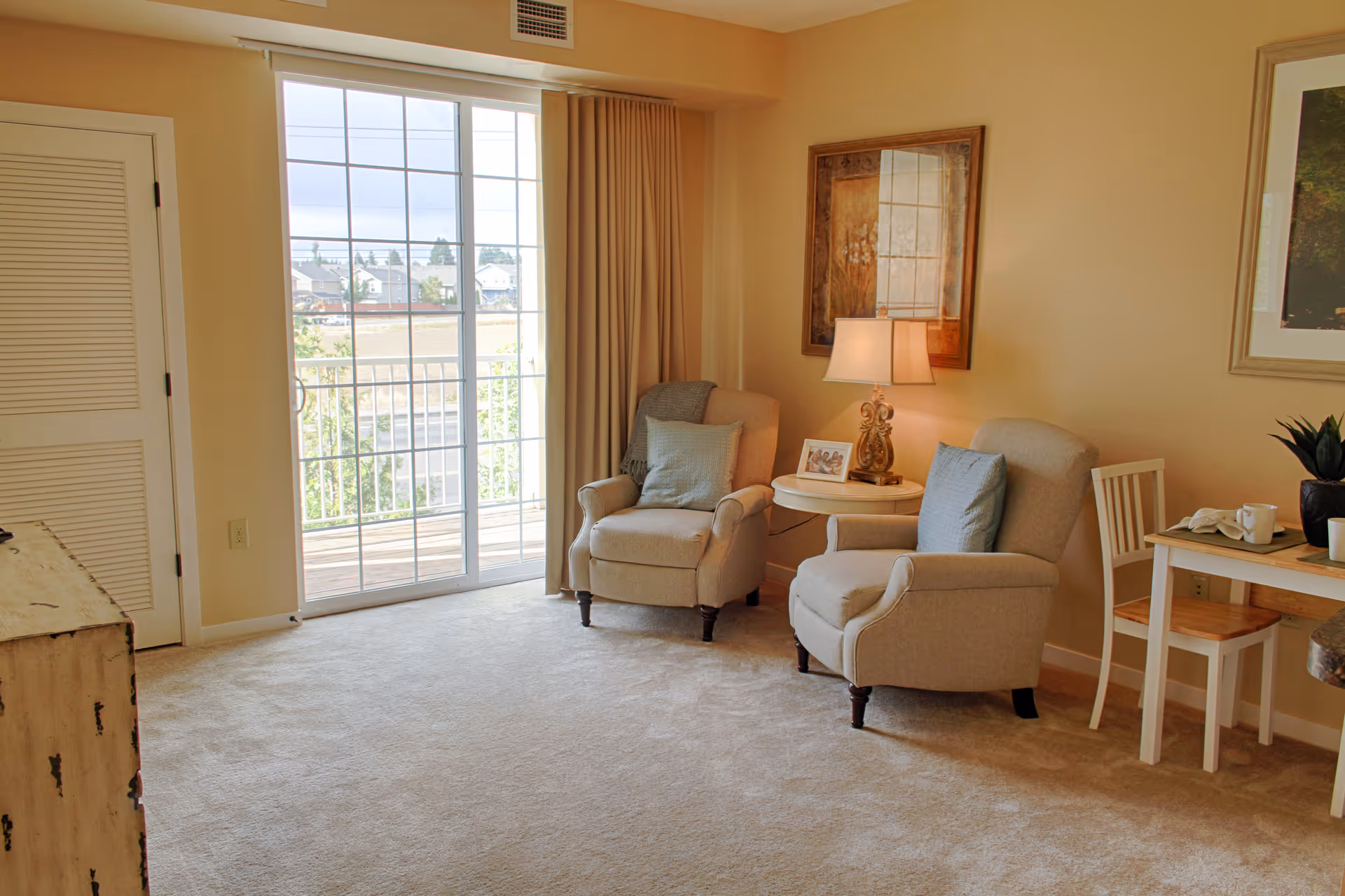 A cozy living room area with two beige armchairs, each with a light blue pillow, positioned around a small round table with a decorative lamp and a framed photo. There is a sliding glass door leading to a balcony with a view of houses and trees outside. A wooden table with two chairs and a plant is visible on the right side. The room has beige walls and carpeted floor.
