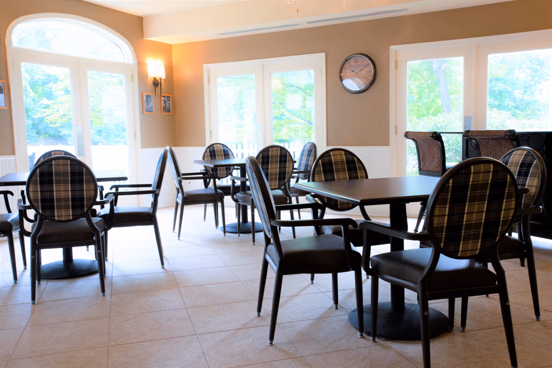 A bright dining room with several tables and chairs featuring plaid upholstery. Large windows and glass doors allow natural light to fill the room, and a clock is mounted on the wall above the doors.