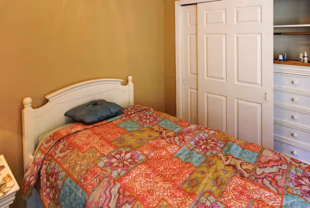 A small bedroom with a single bed covered in a colorful patchwork quilt. The bed has a white wooden headboard and a gray pillow. To the right, there is a closet with sliding white doors, one of which is partially open revealing shelves and drawers inside. The walls are painted beige.