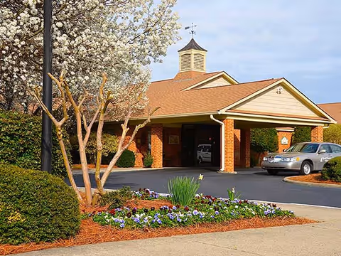 Entrance of a retirement village building with a covered driveway, surrounded by landscaped bushes, flowering plants, and a tree with white blossoms. A silver car is parked near the entrance under a clear sky.