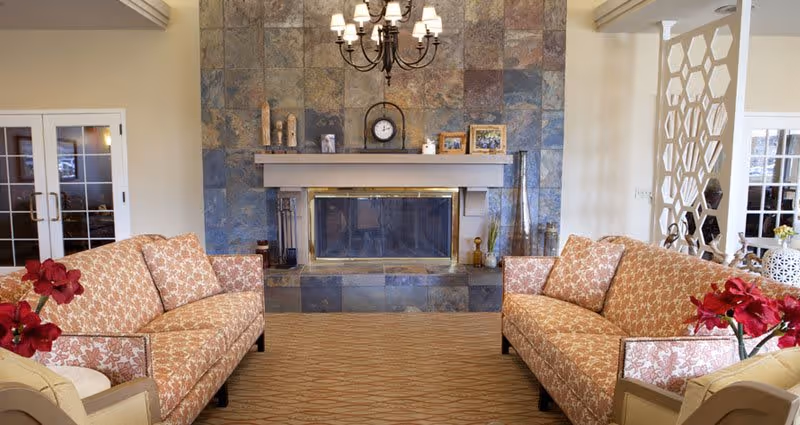 A cozy living room with two patterned sofas facing each other across a carpeted floor. The room features a large stone fireplace with a mantel decorated with candles, a clock, and framed photos. A chandelier hangs above the fireplace, and there are red flowers on side tables next to the sofas. The walls are light-colored, and there are glass doors and decorative partitions in the background.