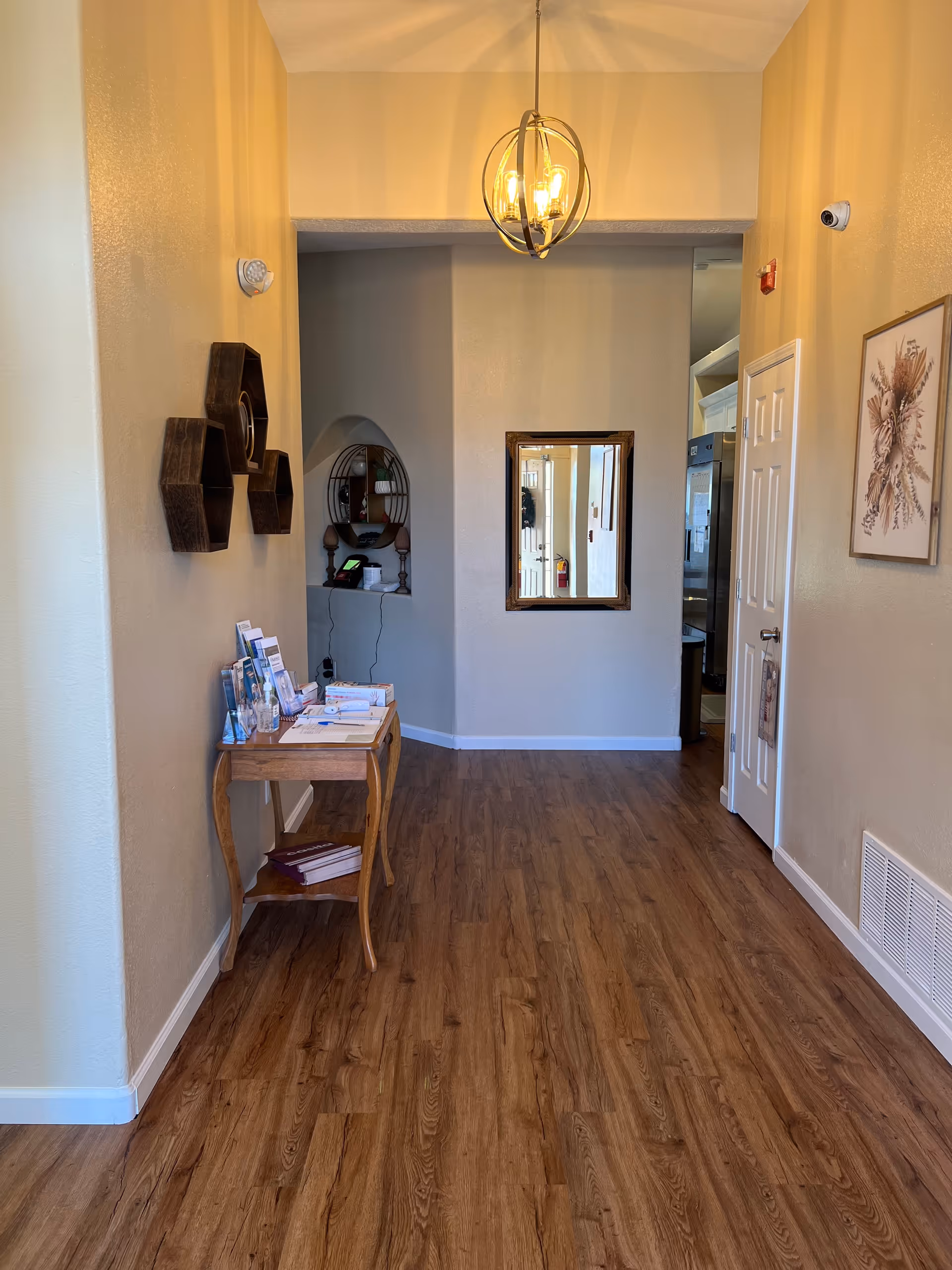 A hallway with wooden flooring and beige walls featuring a small wooden table with brochures and books on the left side. Above the table are three hexagonal wall shelves. A round mirror with decorative items is visible in a recessed wall niche at the end of the hallway. A rectangular mirror hangs on the far wall, reflecting part of the hallway and a door. A modern chandelier with exposed bulbs hangs from the ceiling. On the right wall, there is a framed botanical artwork, a white door, a vent, and a security camera.