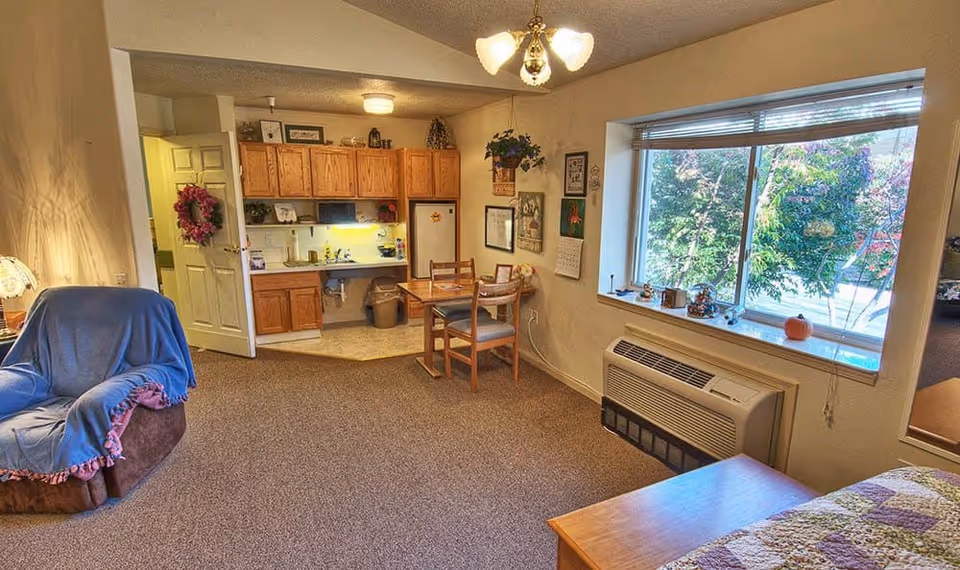 Interior view of a cozy assisted living room with a small kitchenette in the background. The room features a comfortable armchair covered with a blue throw, a wooden dining table with two chairs, a large window with a view of greenery outside, and a heating/cooling unit beneath the window. The kitchenette has wooden cabinets, a sink, and a refrigerator. The walls are decorated with framed pictures and a calendar.