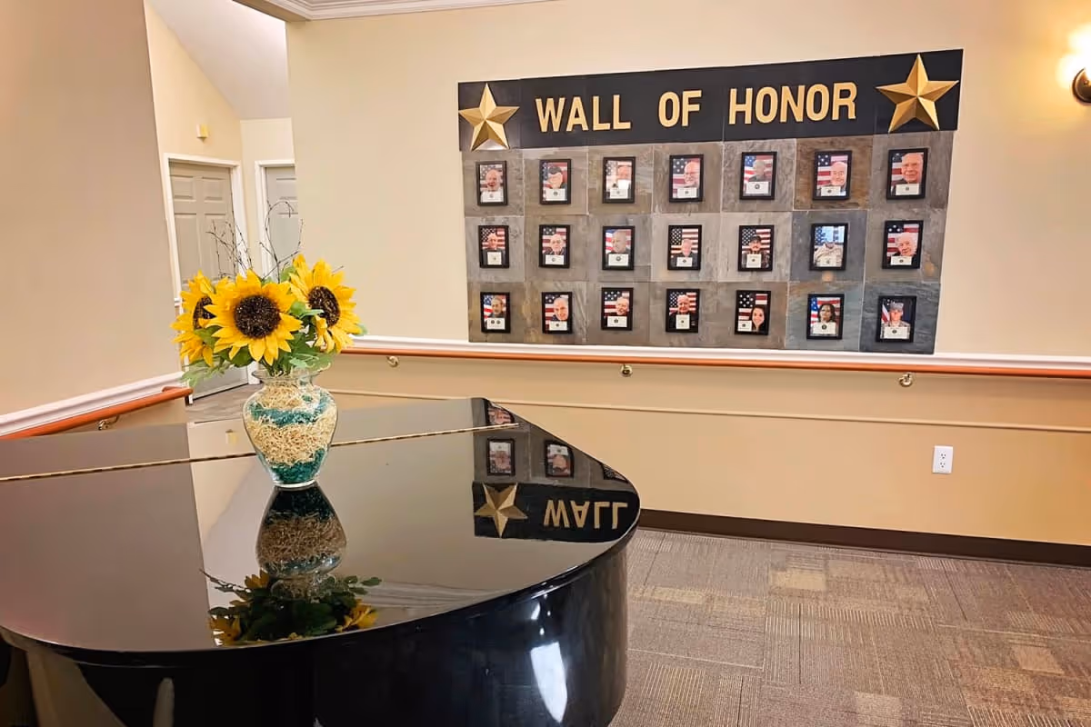 Interior view of a senior living facility hallway featuring a black grand piano with a vase of sunflowers on top. On the wall behind the piano is a 'Wall of Honor' display with framed photos and American flags, decorated with two gold stars.