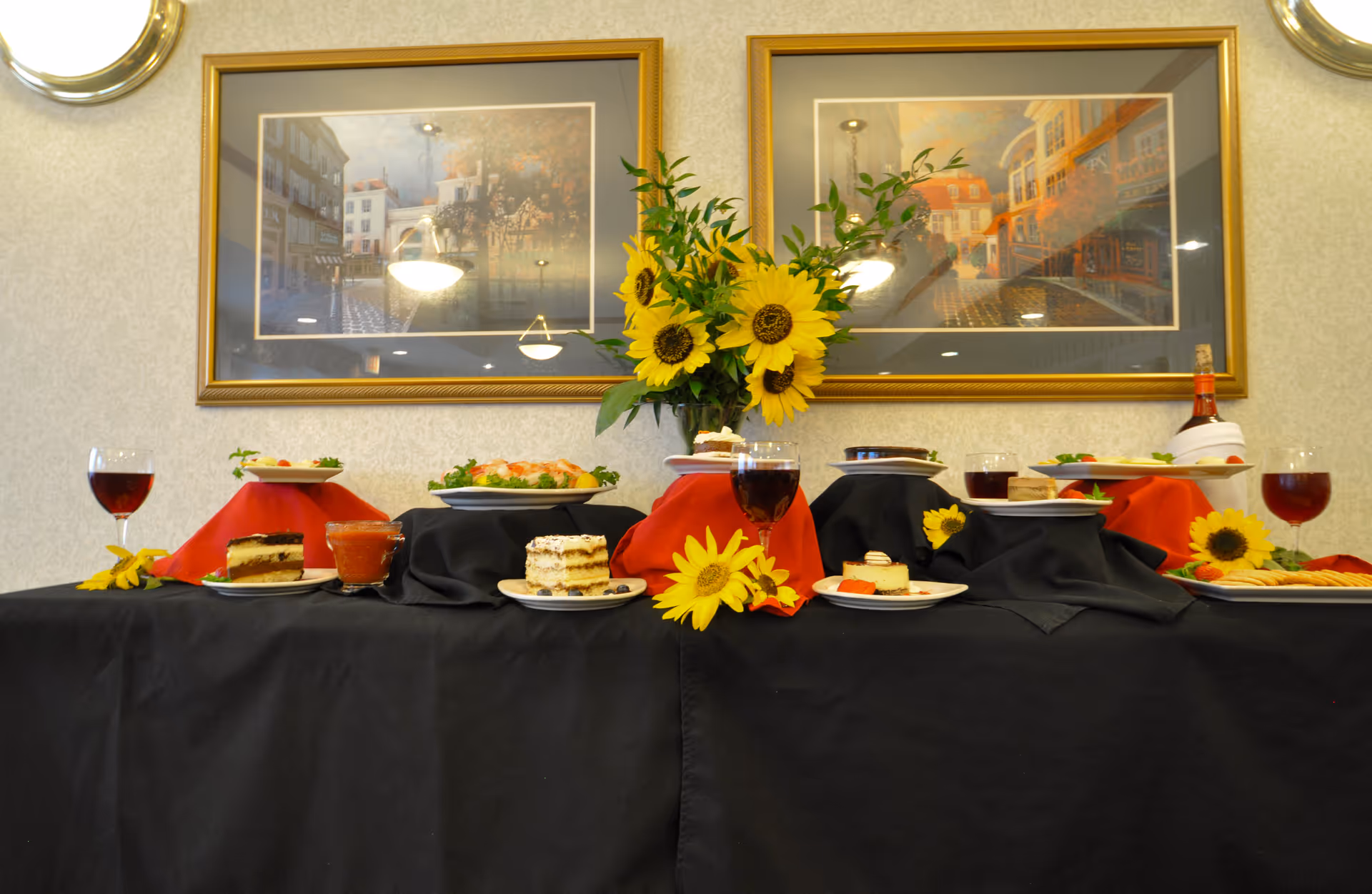 A dining table set with various plated desserts, glasses of red wine, and a centerpiece of bright yellow sunflowers. The table is covered with black and red cloths, and two framed paintings hang on the wall behind the table.