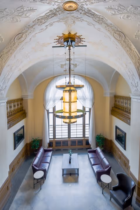 A high-ceilinged elegant lounge area with ornate white plasterwork on the arched ceiling and a large decorative hanging light fixture. The room features two maroon leather sofas facing each other with a rectangular coffee table in between, two small round side tables, a black armchair, and potted plants near large windows with sheer white curtains.