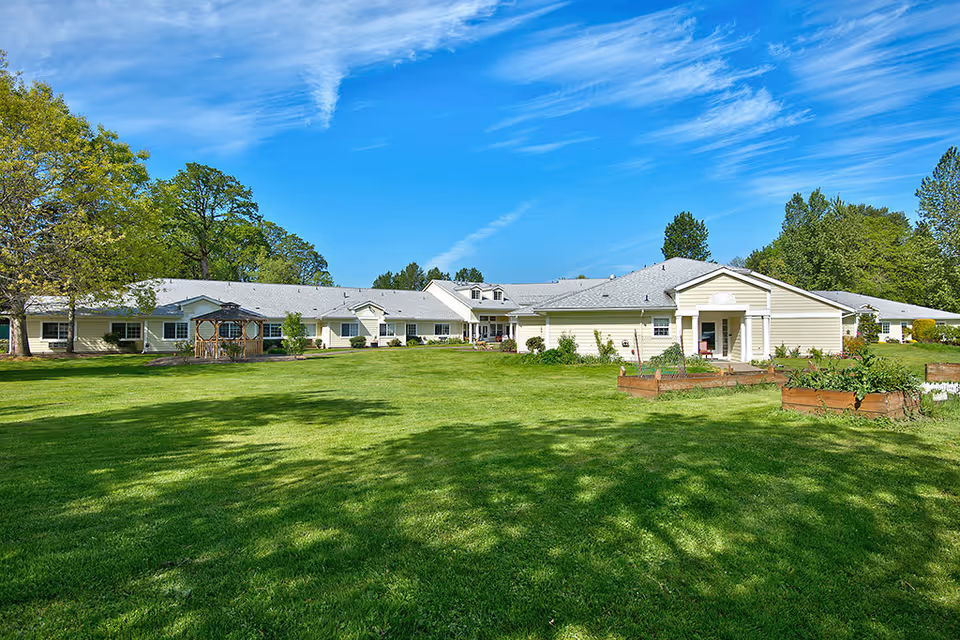 Wide exterior view of Deerfield Village Assisted Living facility showing a single-story building with light-colored siding and a gray roof, surrounded by a large green lawn, trees, and a clear blue sky with wispy clouds.